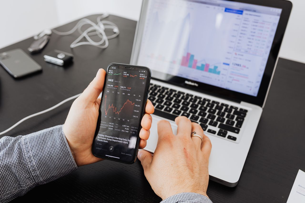 A person reviews stock data on a smartphone and laptop indoors.